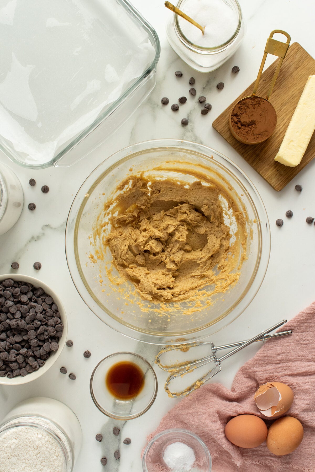 A glass bowl with cookie dough sits on a marble counter surrounded by baking ingredients: chocolate chips, eggs, butter, cocoa powder, sugar, vanilla, flour, a mixer, and a glass baking dish.