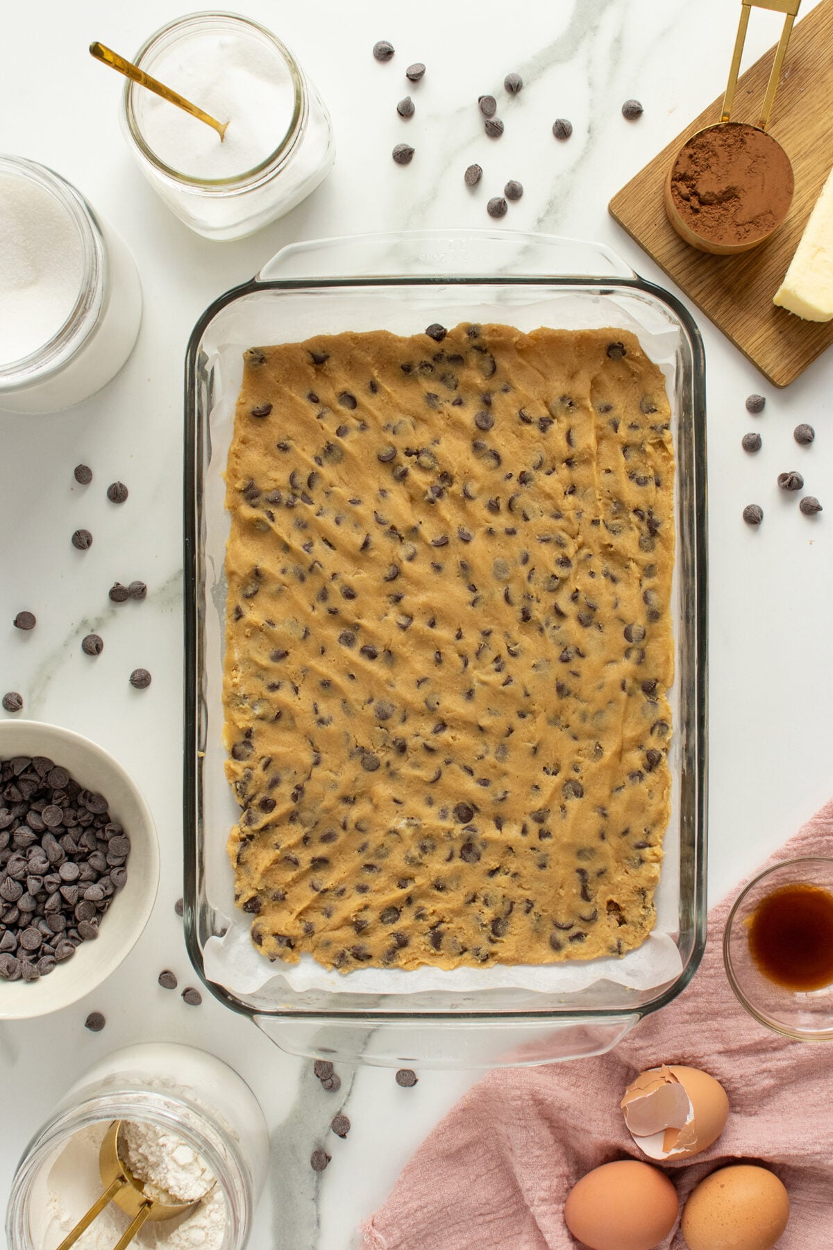 A glass baking dish with chocolate chip cookie dough is surrounded by baking ingredients: chocolate chips, eggs, vanilla extract, sugar, cocoa powder, flour, and butter on a white marble surface.