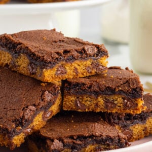 A stack of chocolate and cookie bars with visible chocolate chips, arranged on a pink plate, with a blurred plate of more bars and a glass of milk in the background.