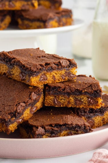 A stack of chocolate and cookie bars with visible chocolate chips, arranged on a pink plate, with a blurred plate of more bars and a glass of milk in the background.