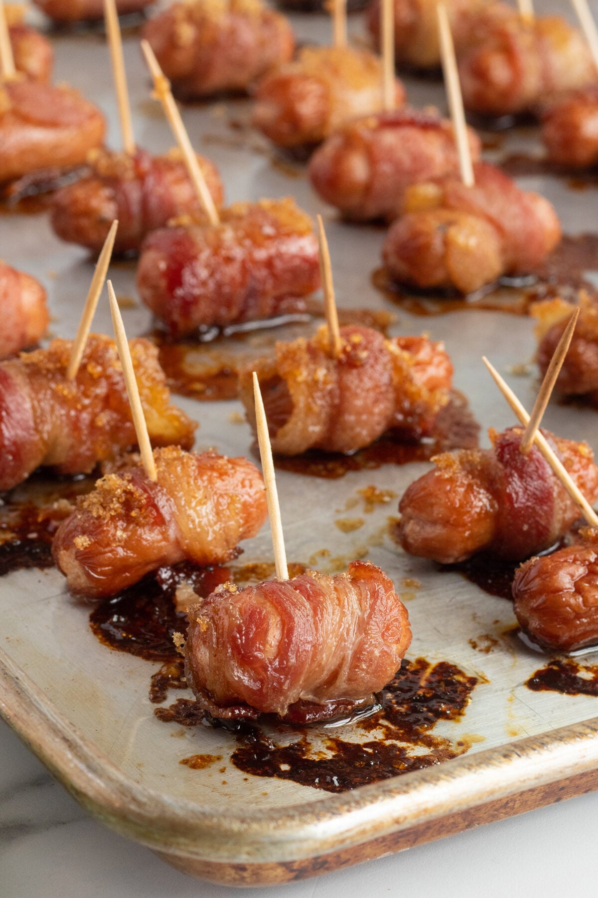 Bacon-wrapped smokies secured with toothpicks sit on a baking sheet, coated in a caramelized glaze. The bacon is browned and crispy, with syrupy sauce pooled around each piece.