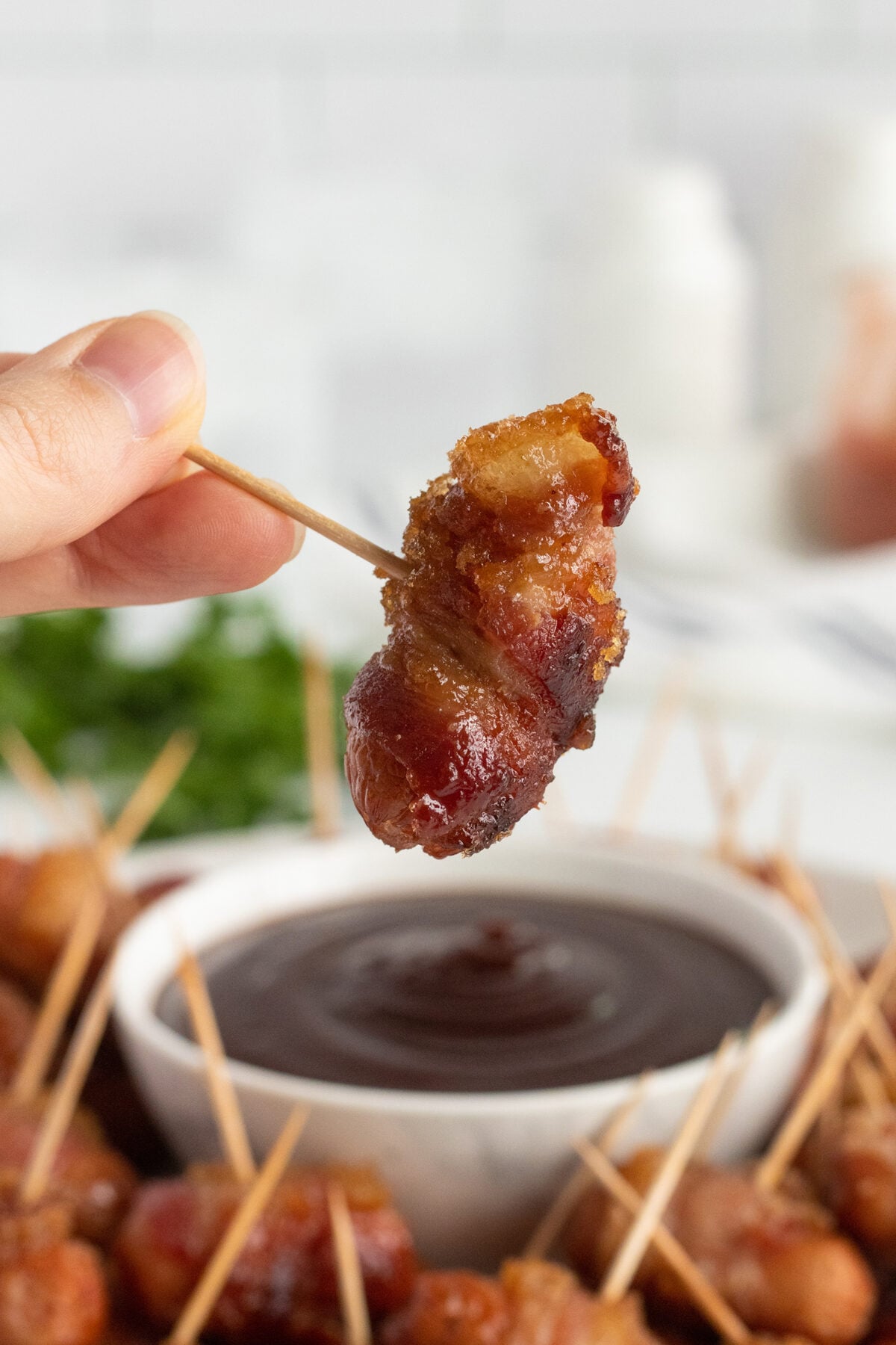 A hand holds a toothpick with a piece of bacon-wrapped food above a bowl of dark dipping sauce, with more bacon-wrapped bites and toothpicks visible in the background.