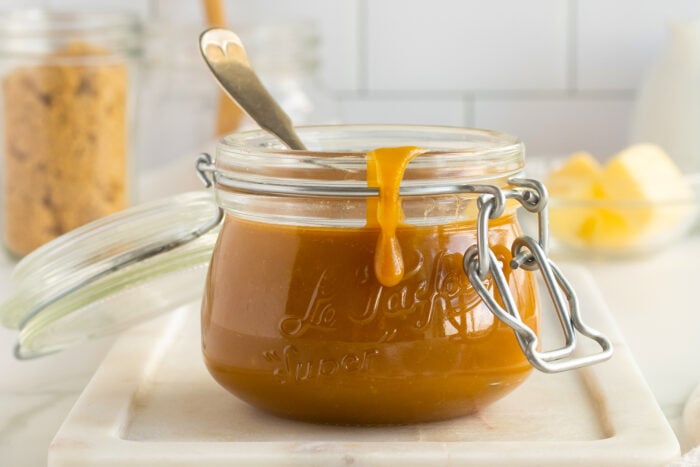 A glass jar filled with caramel sauce, with some sauce dripping down the side and a spoon inside. The jar sits on a marble board, with brown sugar and butter blurred in the background.