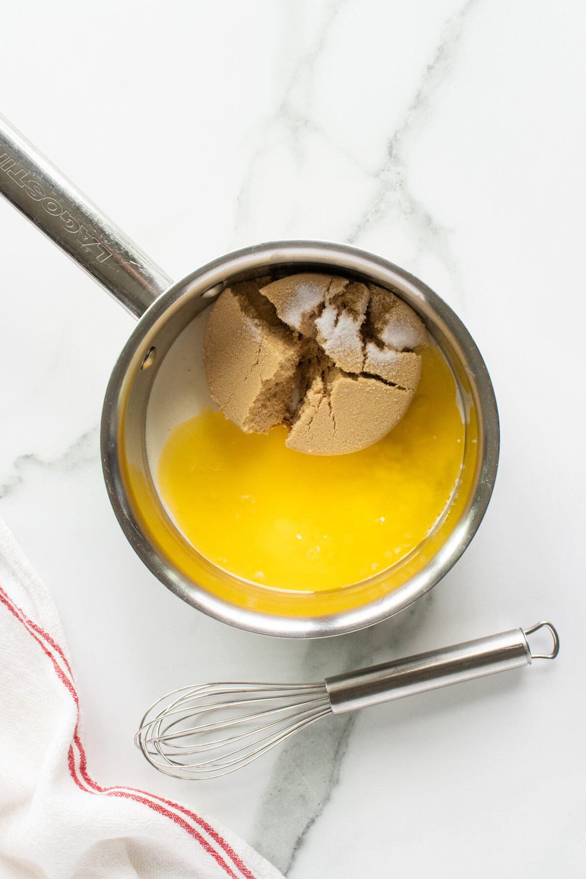 A saucepan containing melted butter and packed brown sugar sits on a marble countertop next to a metal whisk and a white towel with a red stripe.
