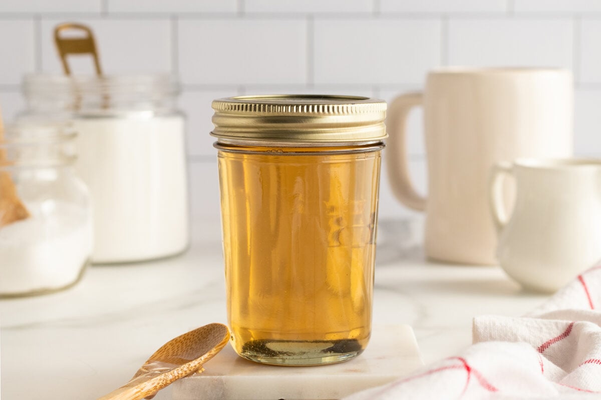 A glass jar filled with golden liquid, sealed with a metal lid, is placed on a marble surface. Nearby are a wooden spoon, a jar of sugar, a white mug, and a white cloth with red stripes.