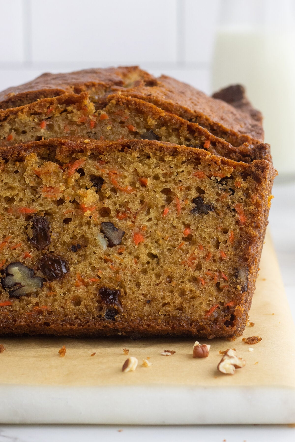 A close-up of a sliced loaf of carrot bread with visible pieces of nuts and raisins, resting on a cutting board with a glass of milk in the blurred background.