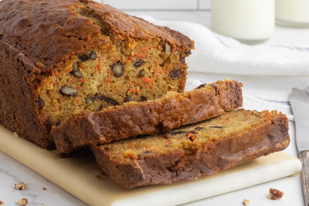 A close-up of a loaf of carrot bread with pecans, partially sliced, resting on parchment paper. The moist, golden interior shows visible carrot shreds and nuts, with a crisp, golden-brown crust.