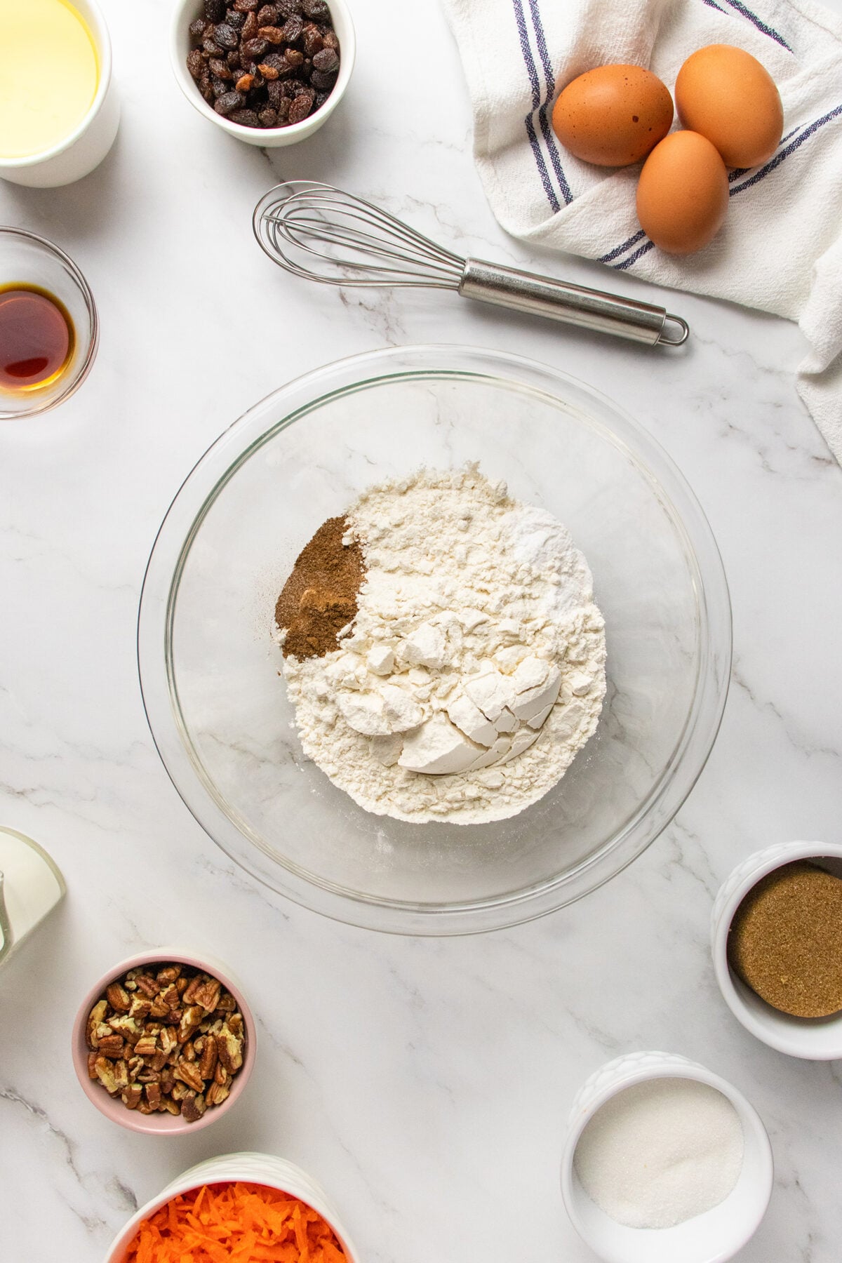 A glass bowl with flour and spices sits on a marble surface, surrounded by eggs, a whisk, a towel, shredded carrots, pecans, raisins, brown sugar, granulated sugar, vanilla, and melted butter.