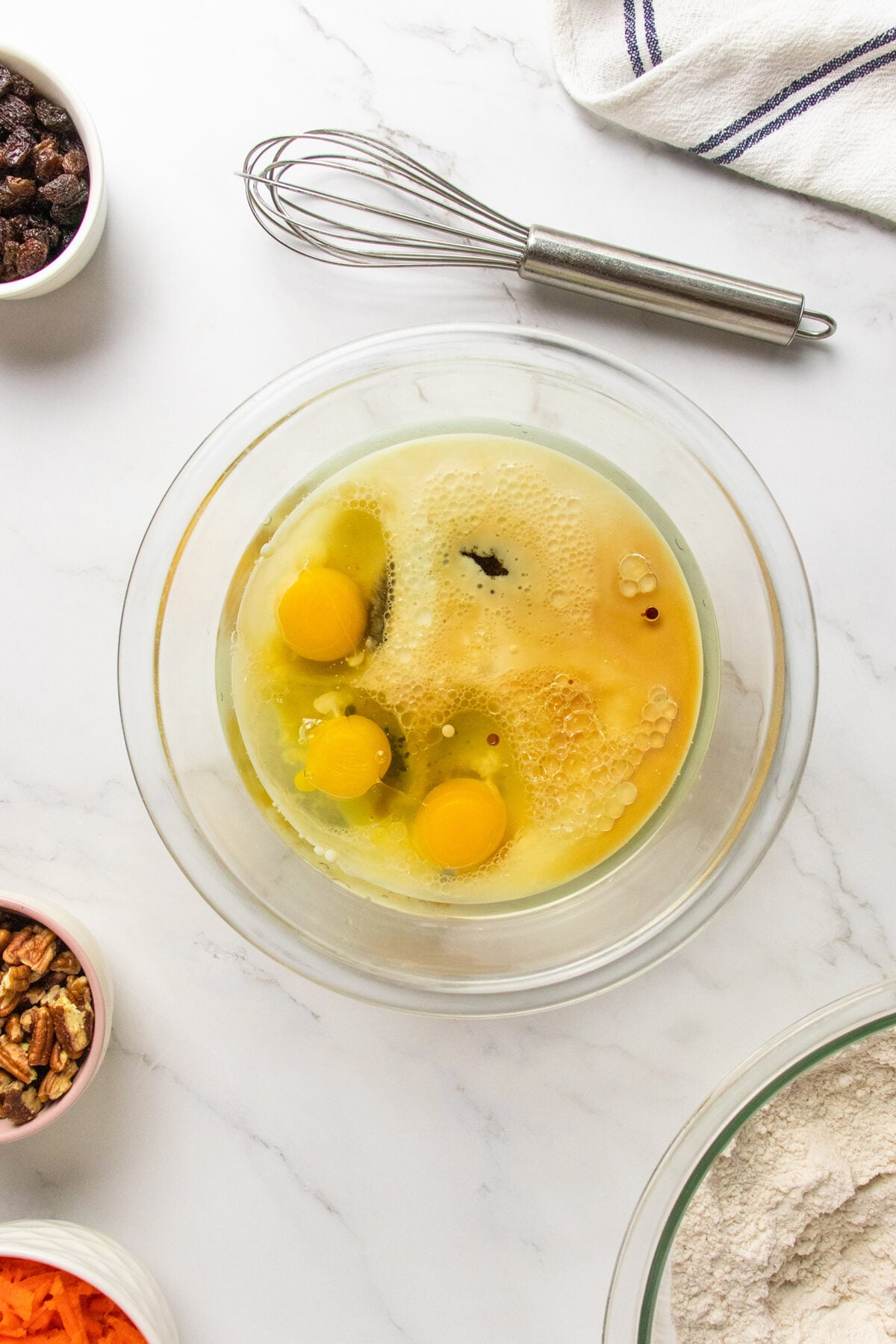 A glass bowl with three cracked eggs, oil, and vanilla extract sits on a white surface surrounded by a whisk, a towel, and bowls of pecans, raisins, shredded carrot, and flour.
