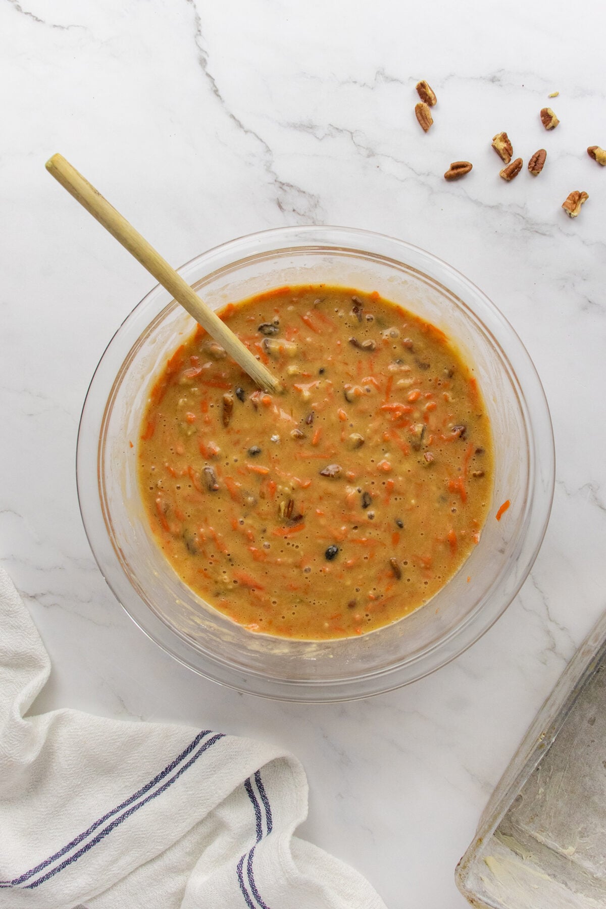 A glass bowl on a marble surface contains carrot cake batter with visible shredded carrots and nuts, being mixed with a wooden spoon. A few chopped pecans are scattered nearby, and a striped towel is partially visible.