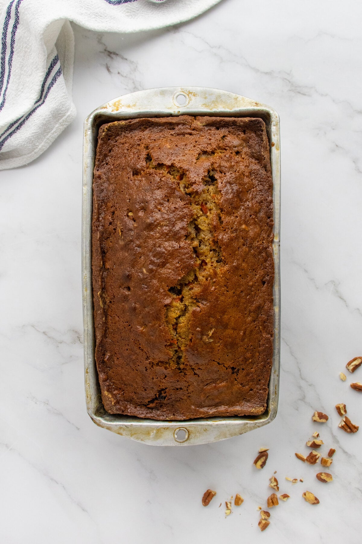 A loaf of banana bread in a metal pan on a marble surface, with a striped towel and chopped pecans nearby.
