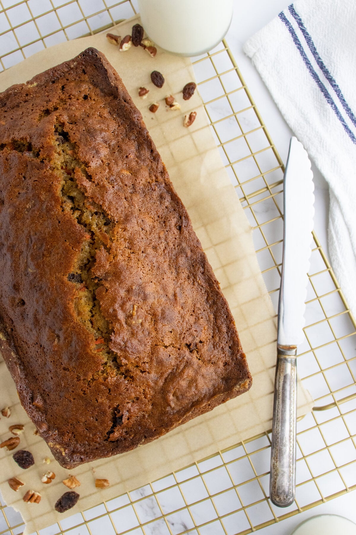 A loaf of banana bread on parchment paper atop a wire cooling rack, with a vintage knife, a white towel, scattered pecans and raisins, and a glass of milk nearby.