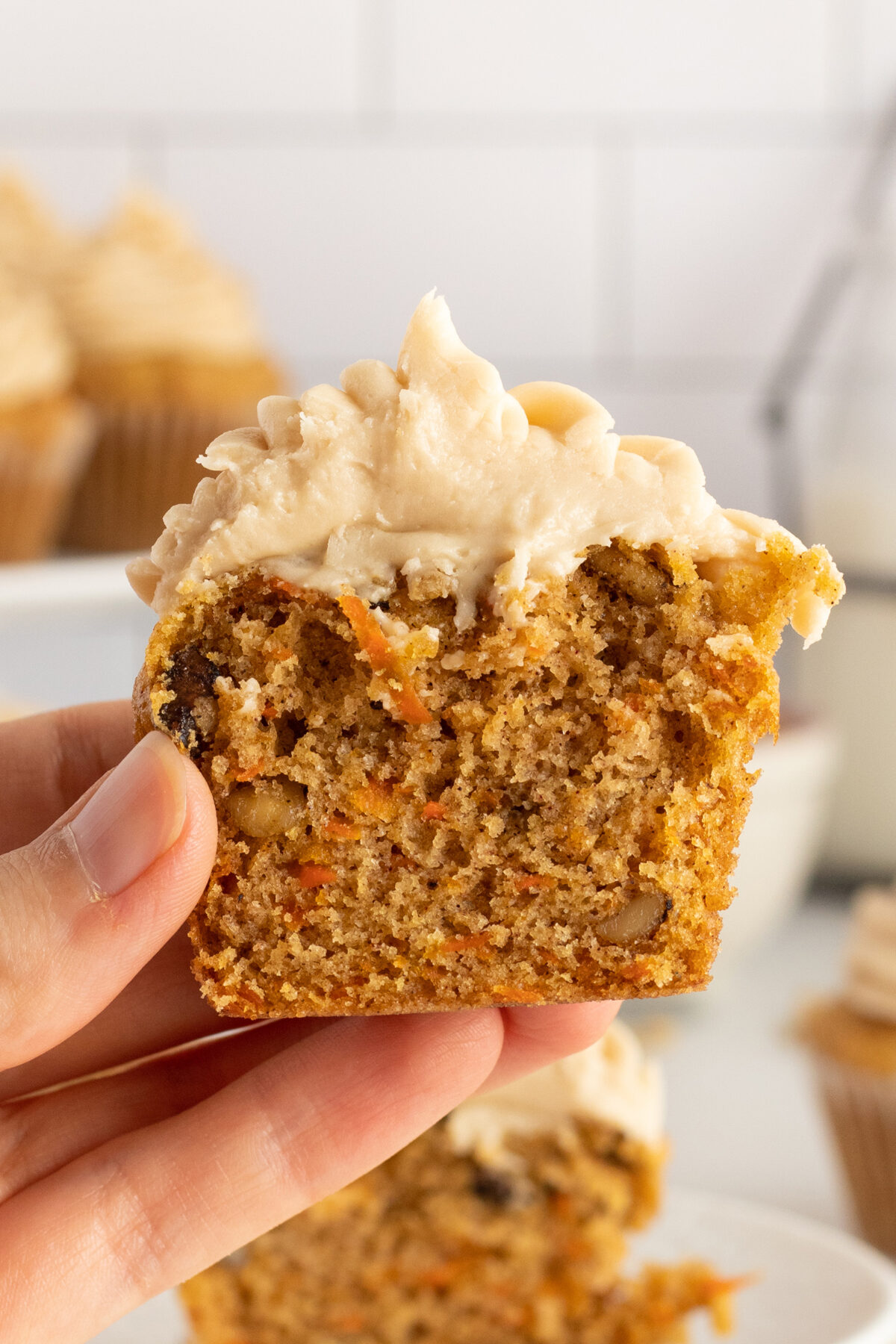 A hand holds a half-eaten carrot cake cupcake topped with creamy frosting. The inside shows bits of carrot and nuts, with other cupcakes blurred in the background.