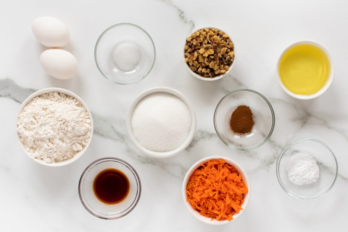 Eleven baking ingredients in bowls on a white marble surface: eggs, flour, sugar, salt, vanilla extract, oil, cinnamon, baking powder, chopped walnuts, and shredded carrots.