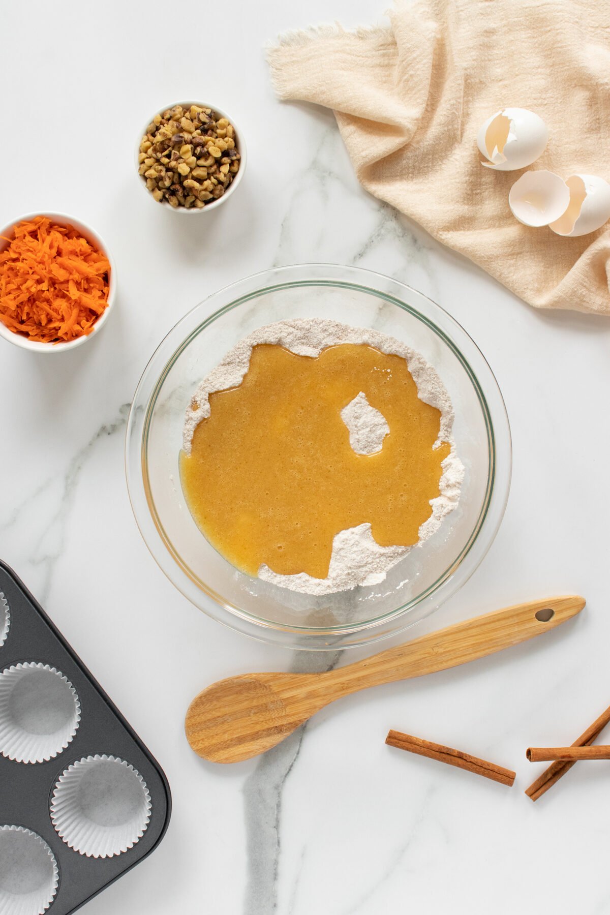 A glass bowl with flour and wet cake batter sits on a marble countertop, surrounded by a muffin tin, a wooden spoon, grated carrots, chopped walnuts, cinnamon sticks, eggshells, and a beige cloth.