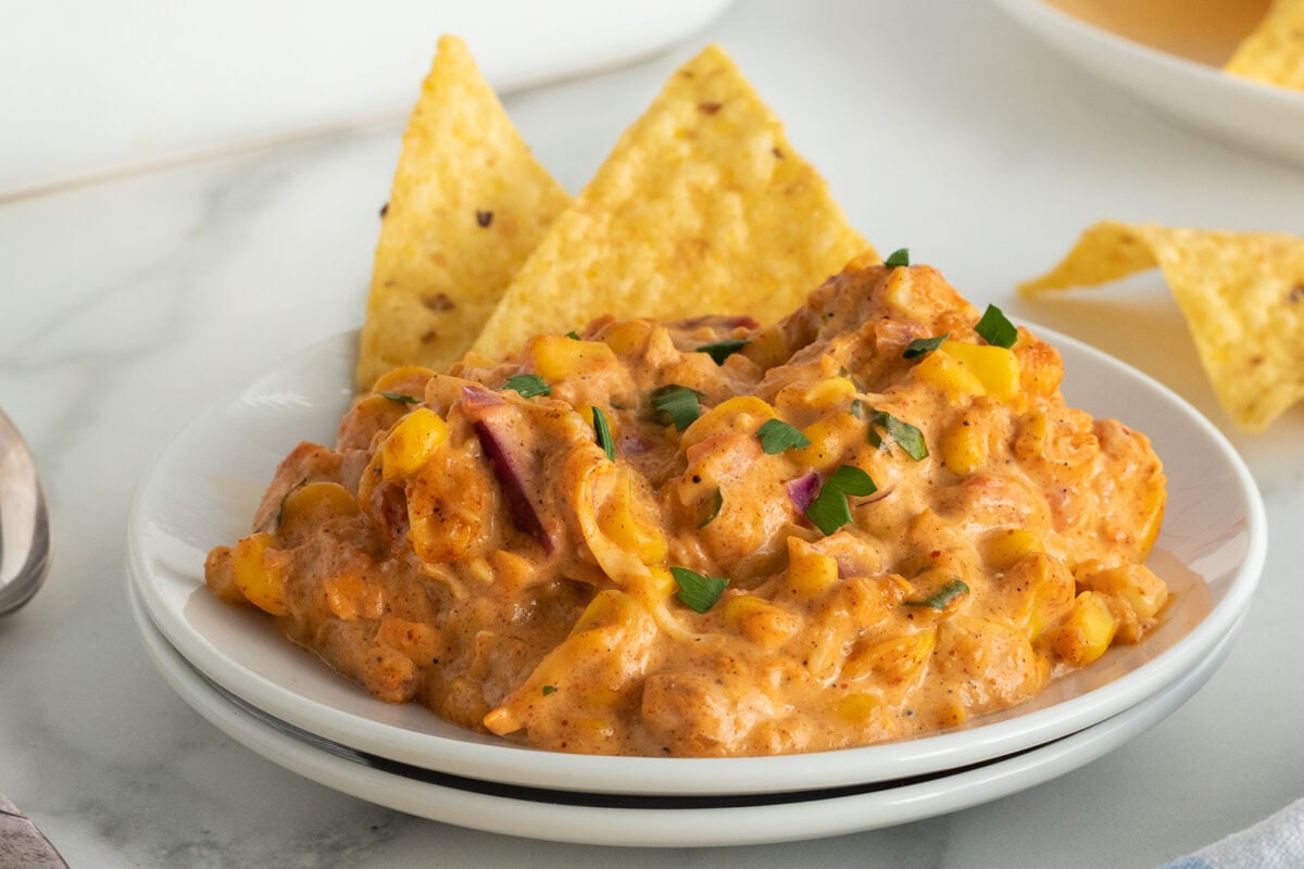 A plate of creamy, cheesy dip with visible corn, diced tomatoes, and herbs, garnished with parsley, served alongside two triangular tortilla chips on a white plate.