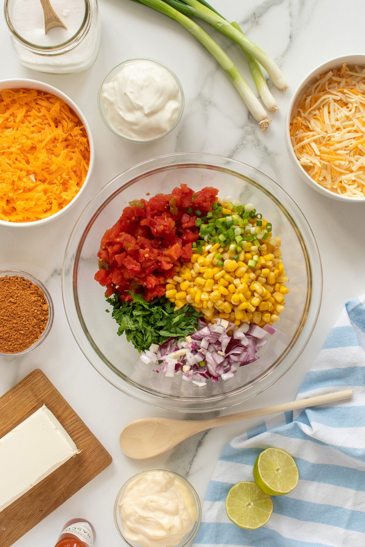 A glass bowl with chopped tomatoes, corn, green onions, cilantro, and red onions, surrounded by shredded cheese, sour cream, cream cheese, limes, taco seasoning, green onions, and a wooden spoon on a marble surface.