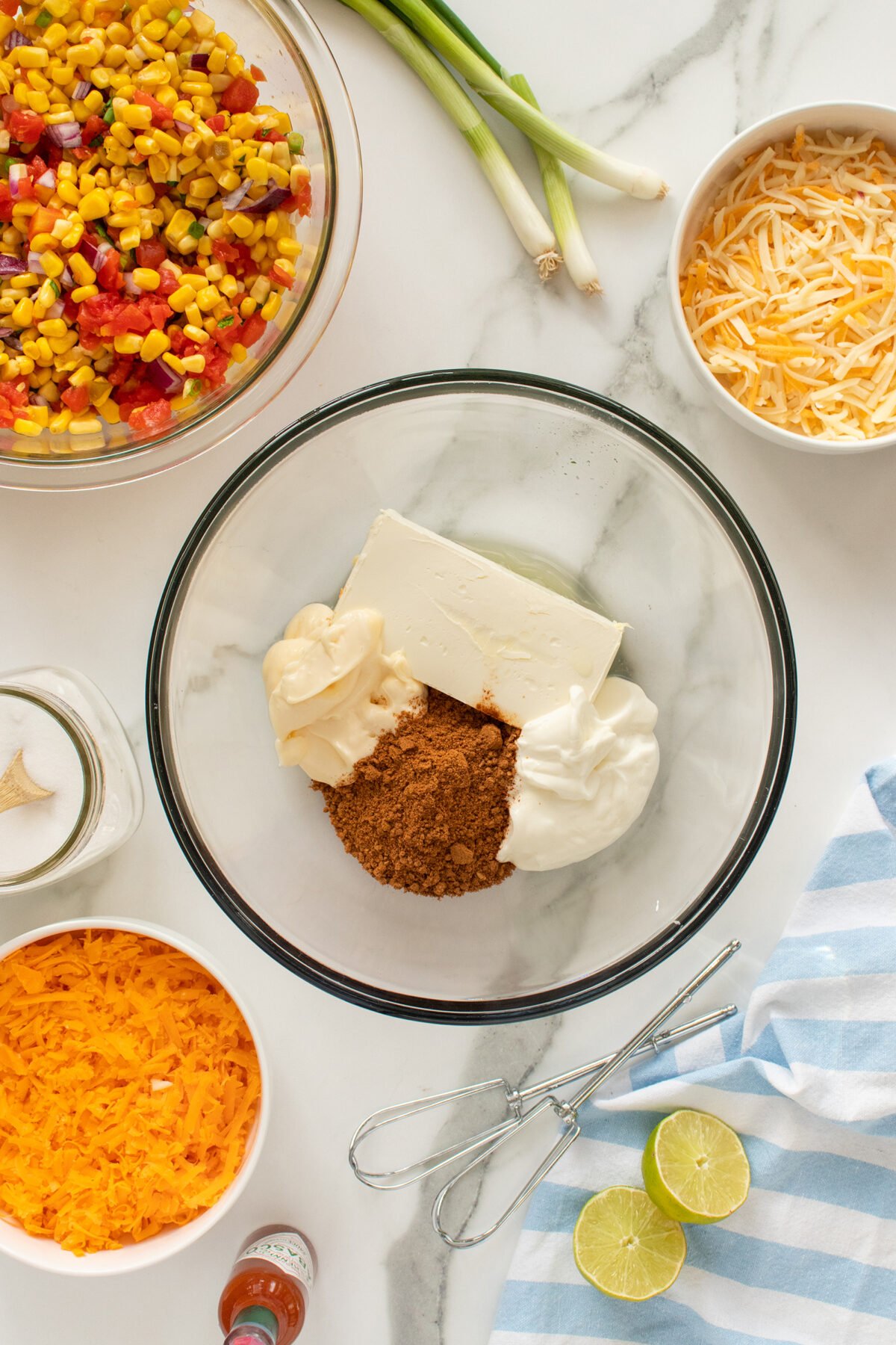 A glass bowl holds cream cheese, mayonnaise, and taco seasoning. Surrounding it are bowls of shredded cheese, corn salsa, green onions, lime halves, a whisk, and a blue-striped towel on a marble surface.