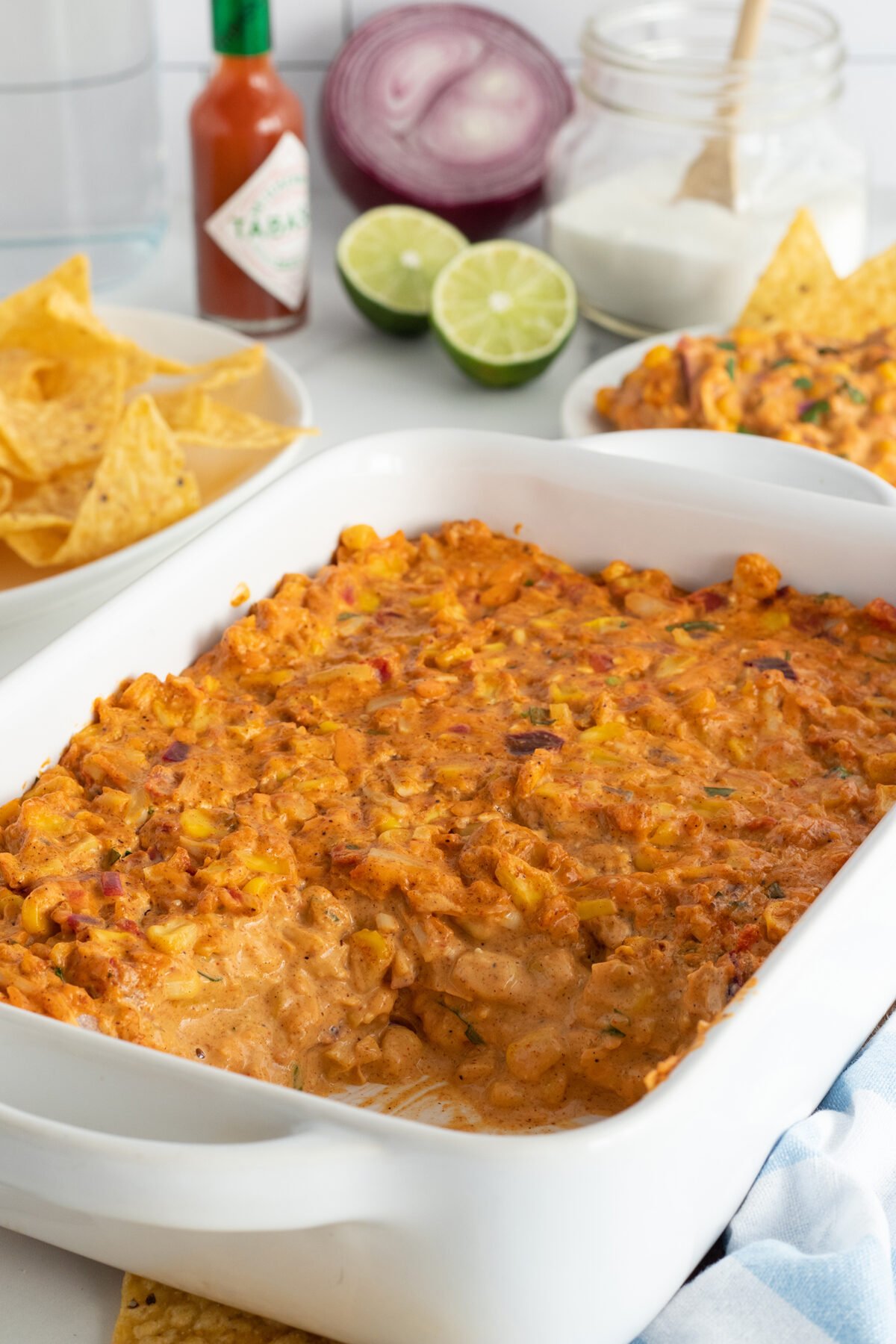 A white casserole dish filled with cheesy corn dip, surrounded by tortilla chips, a halved lime, red onion, a jar of sour cream, and a bottle of Tabasco sauce on a kitchen counter.
