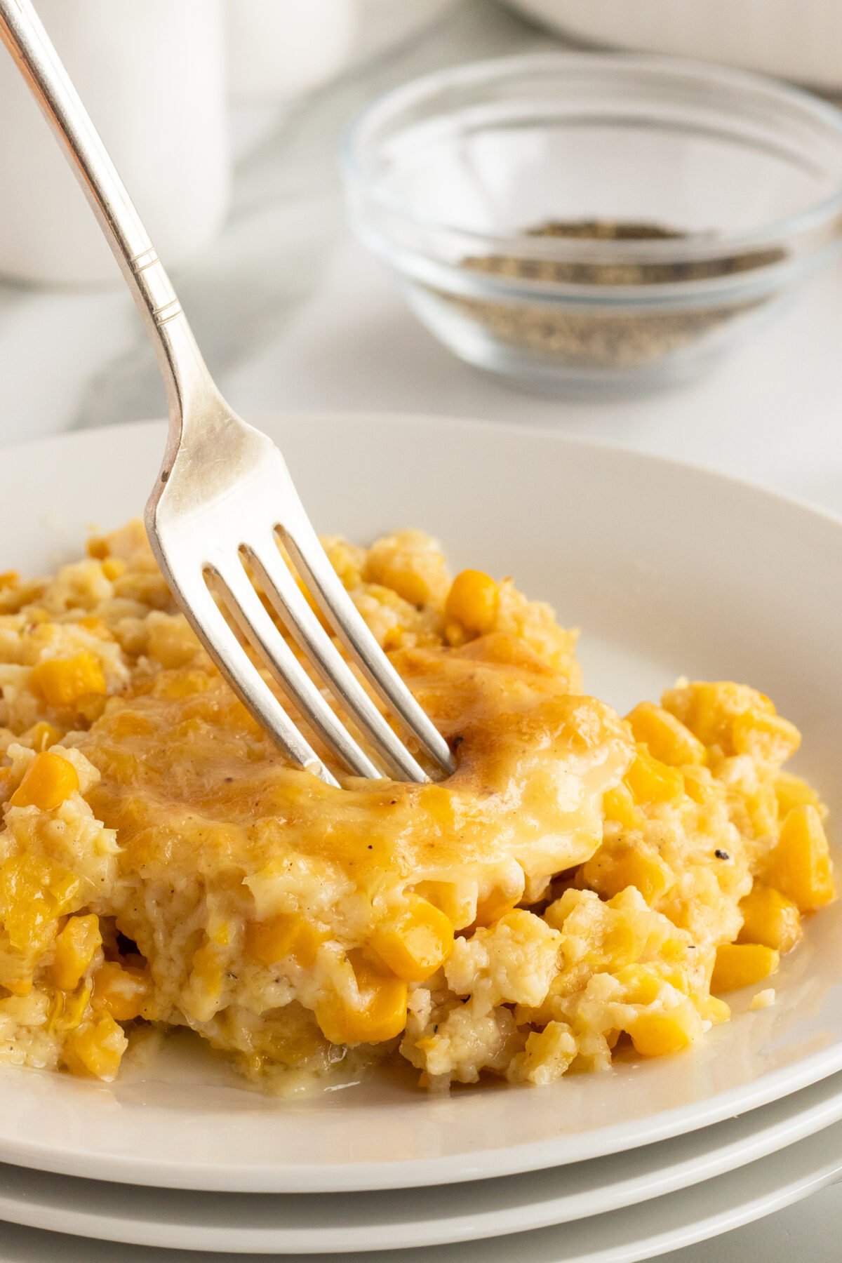 A fork pressing into a serving of creamy corn casserole on a white plate, with visible corn kernels and a golden baked top. A small glass bowl of ground black pepper is in the blurred background.