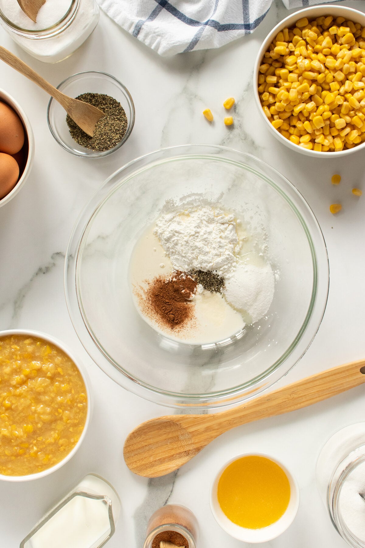 A glass bowl with flour, sugar, and spices sits on a white countertop surrounded by corn, eggs, mashed corn, milk, butter, black pepper, and kitchen utensils.