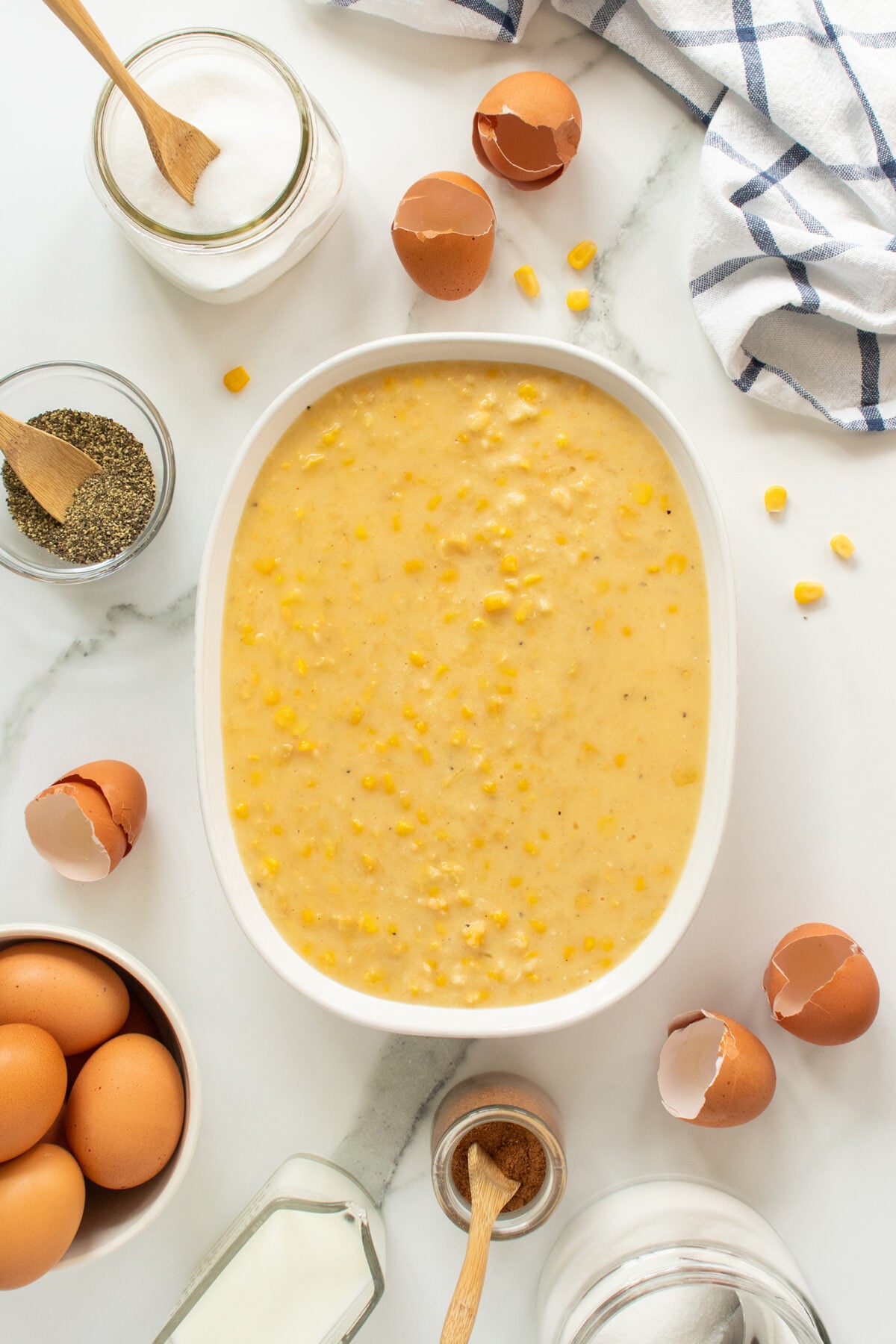 A white baking dish filled with uncooked corn pudding mixture sits on a white marble surface, surrounded by eggs, cracked eggshells, jars of sugar, pepper, cinnamon, and a blue-striped kitchen towel.