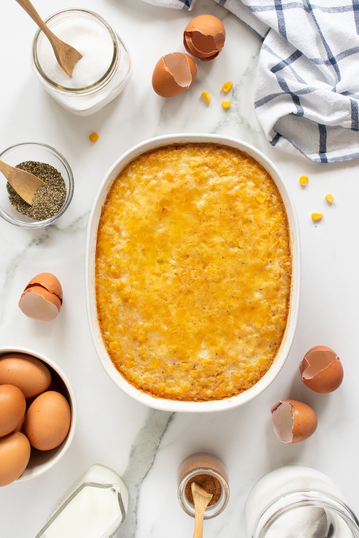 A baked cheesy casserole in a white dish surrounded by eggs, cracked eggshells, pepper, a jar of milk, spices, and a striped kitchen towel on a white marble surface.