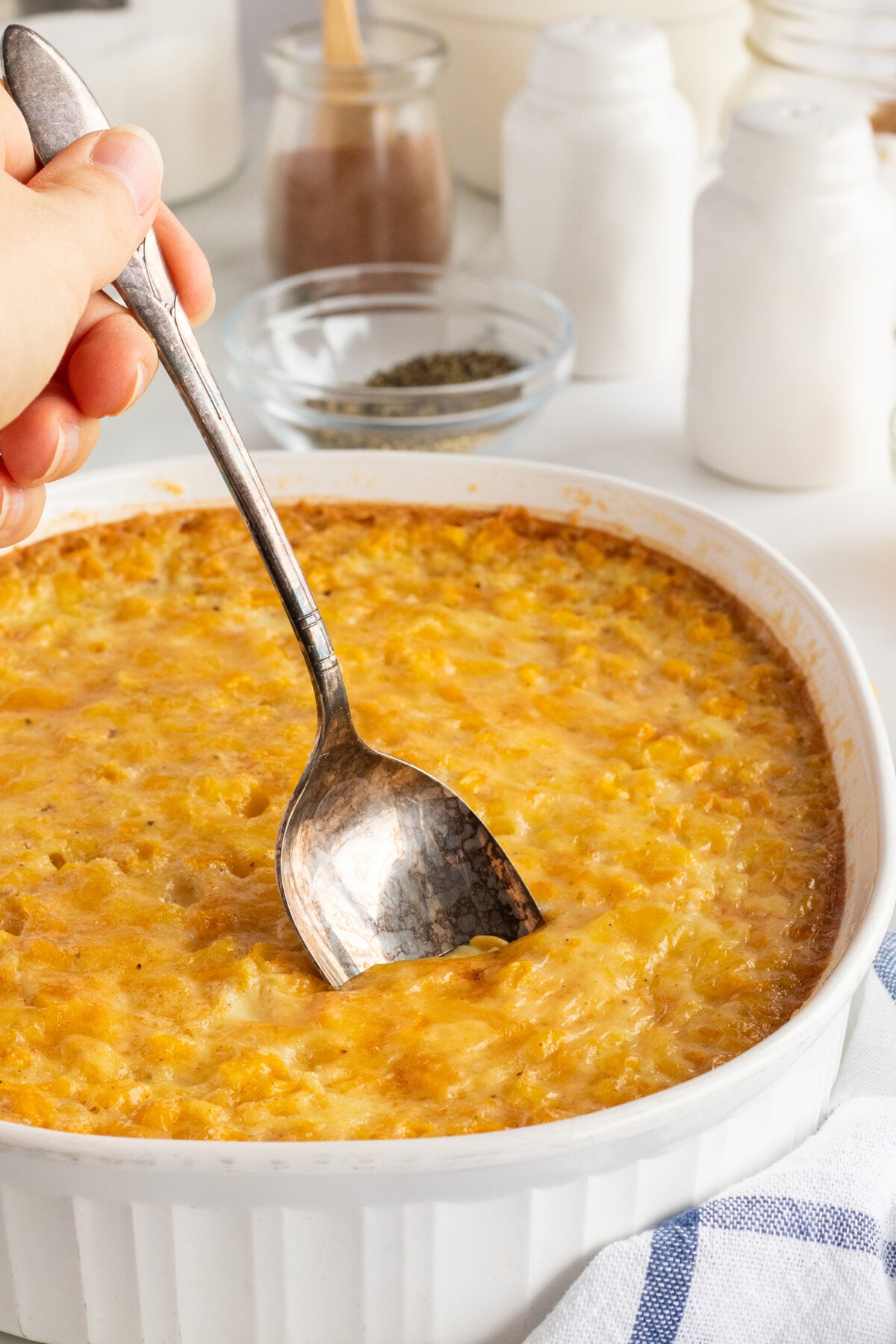 A hand holding a spoon is scooping into a baked cheesy corn casserole in a white dish, surrounded by seasoning jars and a blue-striped kitchen towel on a white surface.
