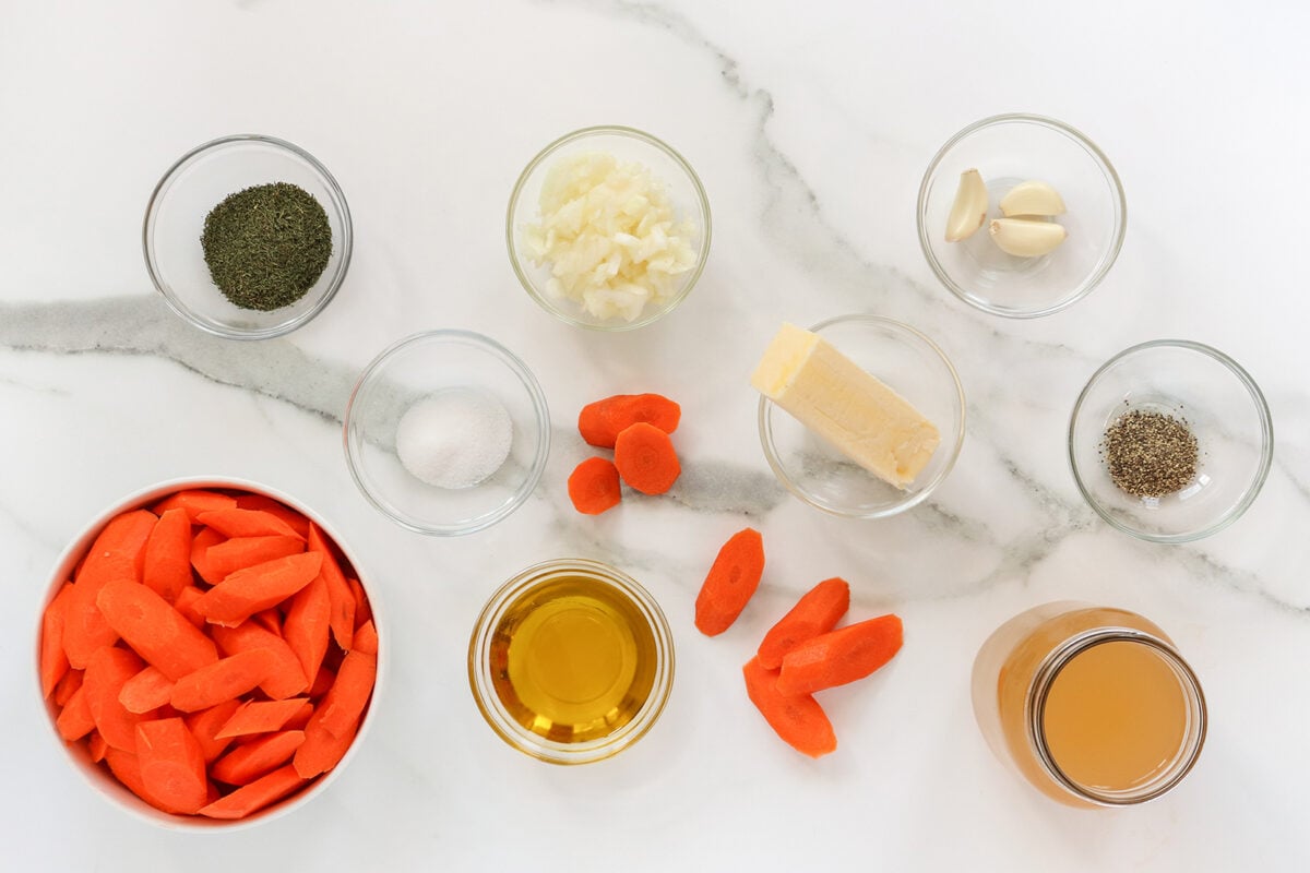 A marble countertop with bowls of ingredients including chopped carrots, olive oil, butter, chopped onions, garlic cloves, dried herbs, salt, pepper, and a jar of broth arranged neatly.