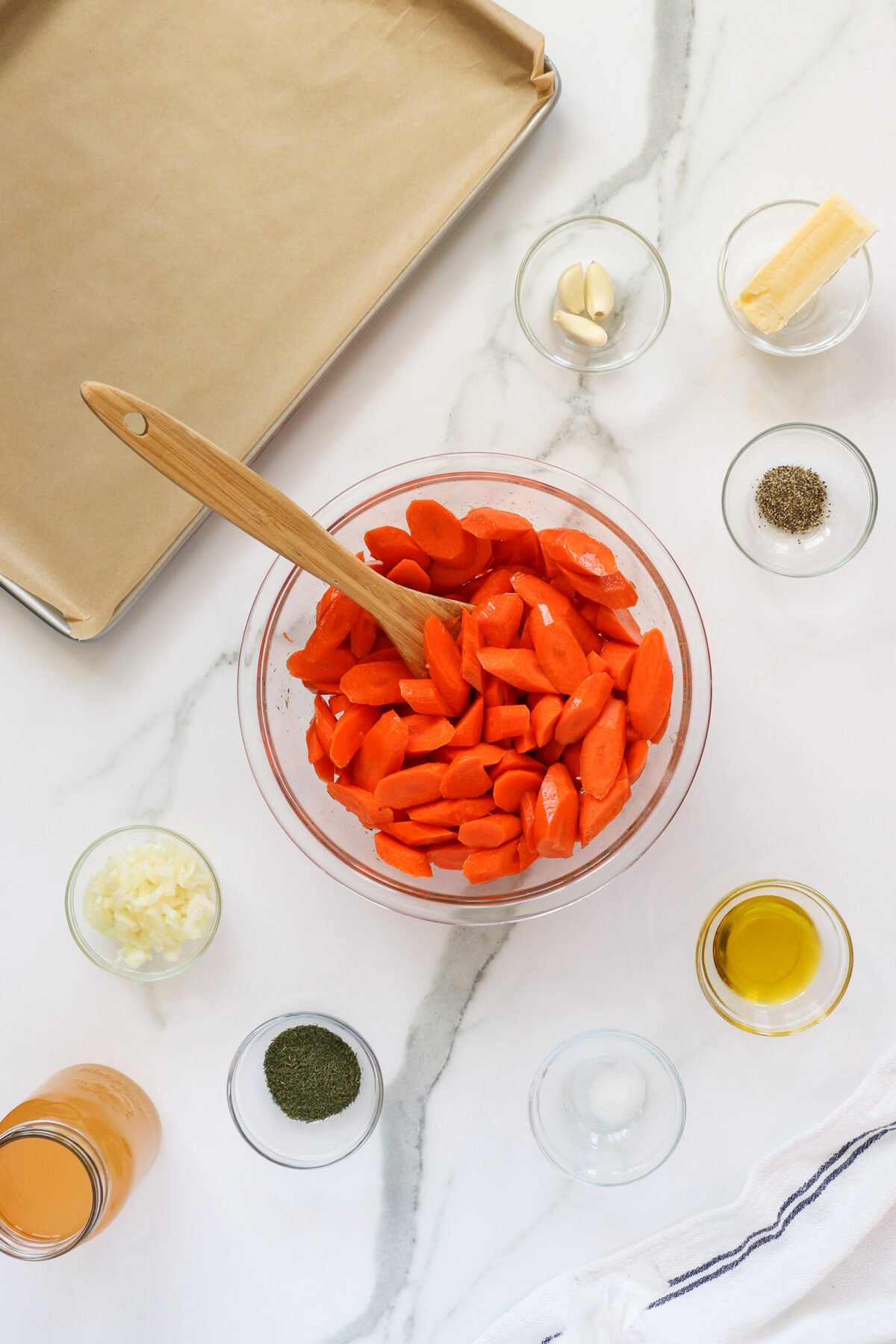 A glass bowl of sliced carrots with a wooden spoon is surrounded by small bowls holding chopped garlic, whole garlic cloves, seasonings, oil, broth, and a baking sheet lined with parchment paper on a marble countertop.