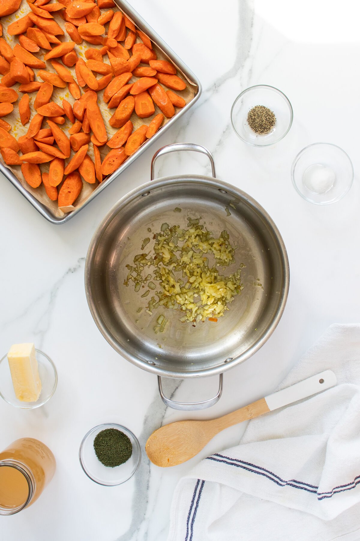 Top-down view of chopped onions sautéing in a pot, surrounded by sliced carrots on a baking tray, bowls of pepper, herbs, butter, broth, and a wooden spoon on a white marble surface.