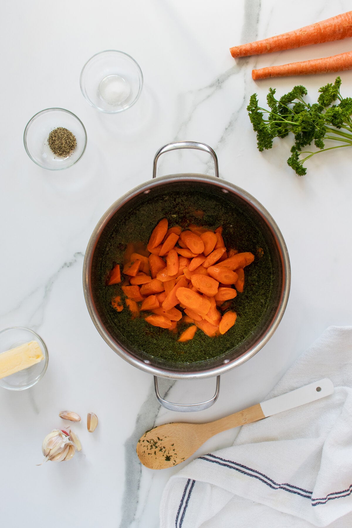 A pot with chopped carrots and herbs sits on a white countertop, surrounded by fresh parsley, whole carrots, a stick of butter, garlic cloves, a wooden spoon, and small bowls of pepper and salt.