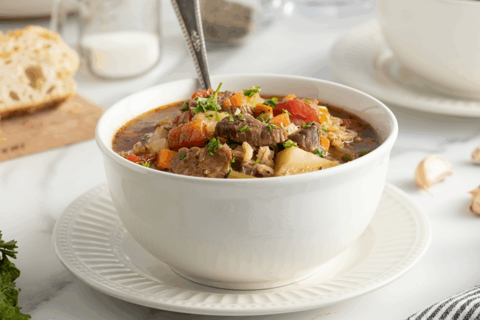A white bowl filled with hearty beef and vegetable soup, garnished with chopped parsley, sits on a matching saucer. A spoon is inside the bowl. In the background, there is bread and a milk jar.