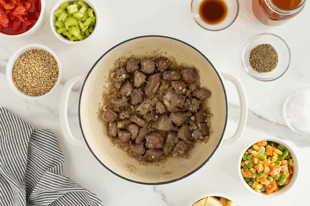 Chunks of beef cooking with onions in a white pot, surrounded by small bowls containing celery, diced tomatoes, mixed vegetables, cubed potatoes, seasonings, broth, barley, and a striped cloth on a white counter.