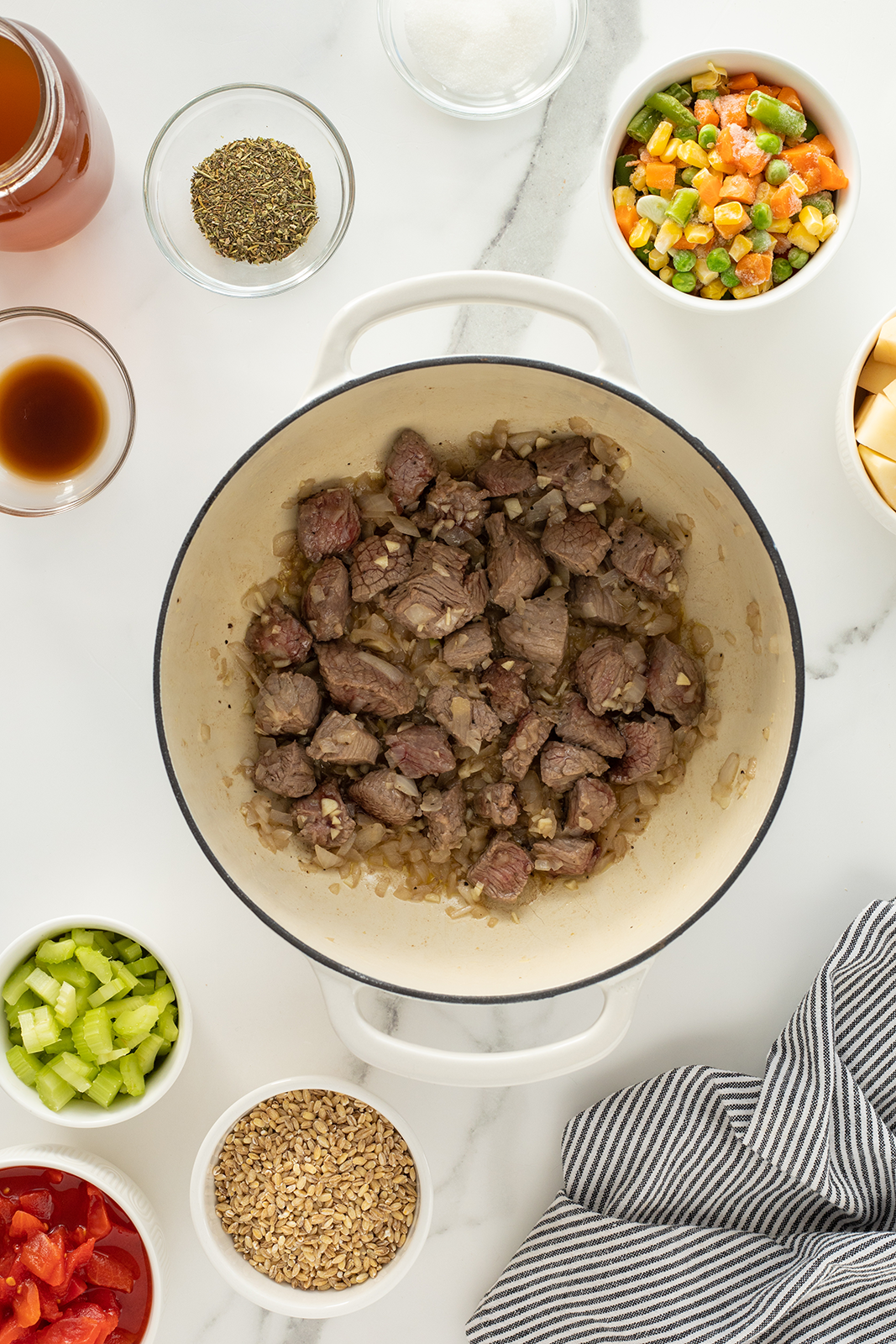 Chunks of beef cooking with onions in a white pot, surrounded by small bowls containing celery, diced tomatoes, mixed vegetables, cubed potatoes, seasonings, broth, barley, and a striped cloth on a white counter.