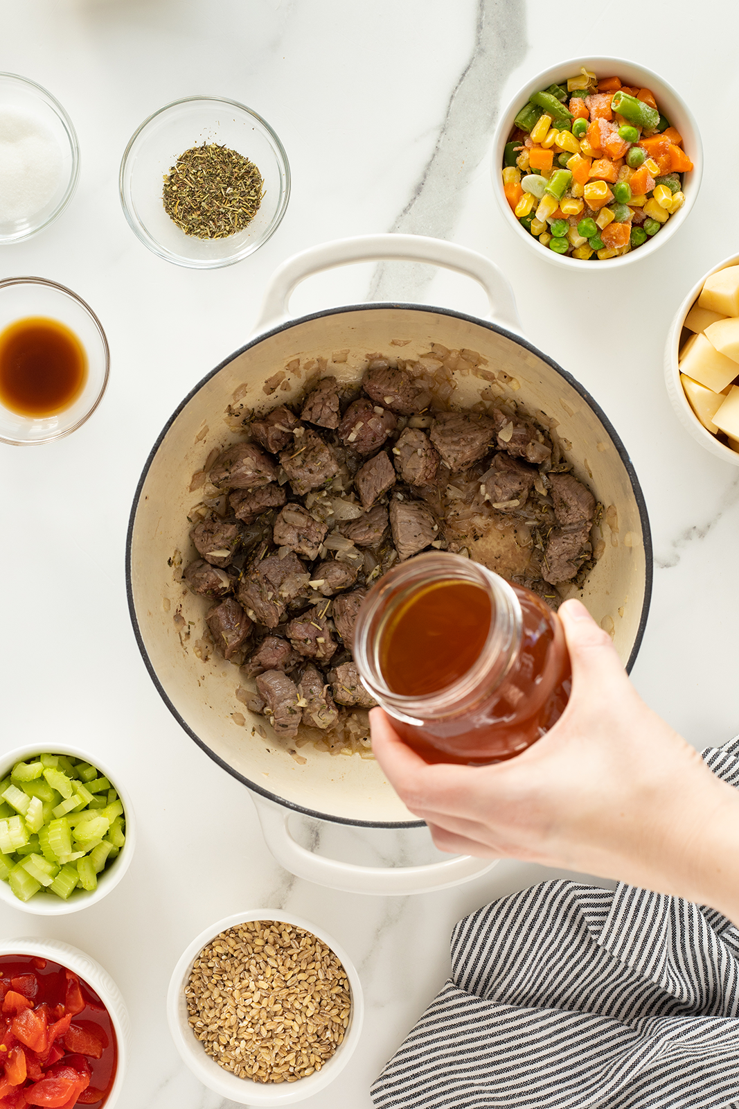 A hand pours broth from a jar into a pot with cooked beef. Around the pot are bowls with celery, mixed vegetables, tomatoes, potatoes, barley, herbs, and seasonings on a white countertop.