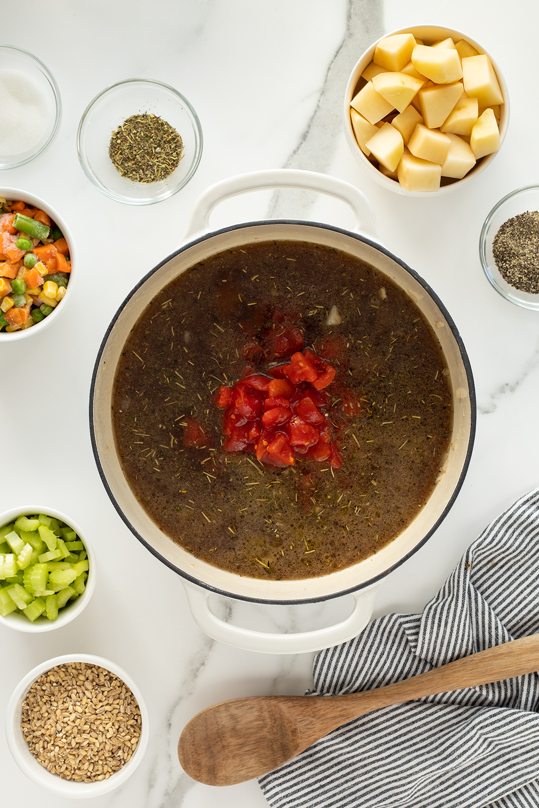 A pot of soup with herbs and diced tomatoes, surrounded by bowls of chopped vegetables, seasonings, grains, and a wooden spoon on a striped cloth, all on a white countertop.