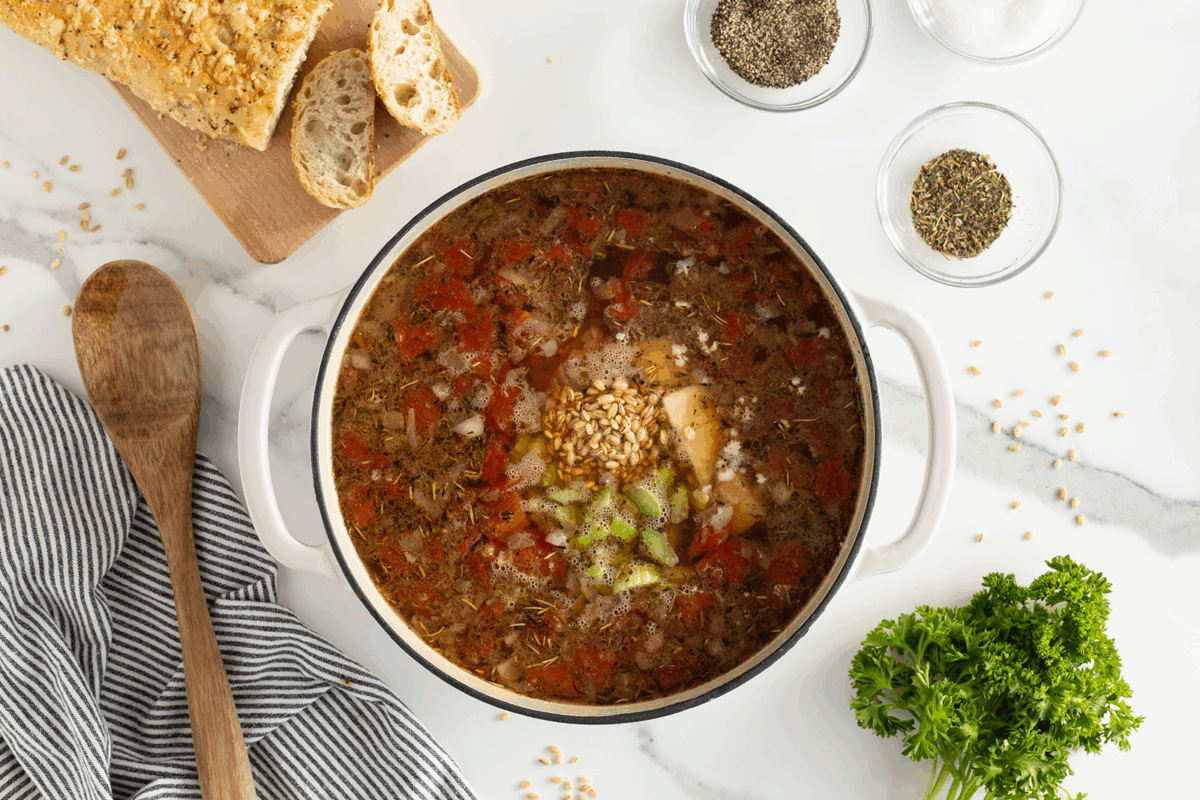 A pot of vegetable and grain soup with chopped tomatoes, celery, and herbs sits on a white counter. Nearby are a wooden spoon, striped towel, sliced bread, bowls of spices, and fresh parsley.
