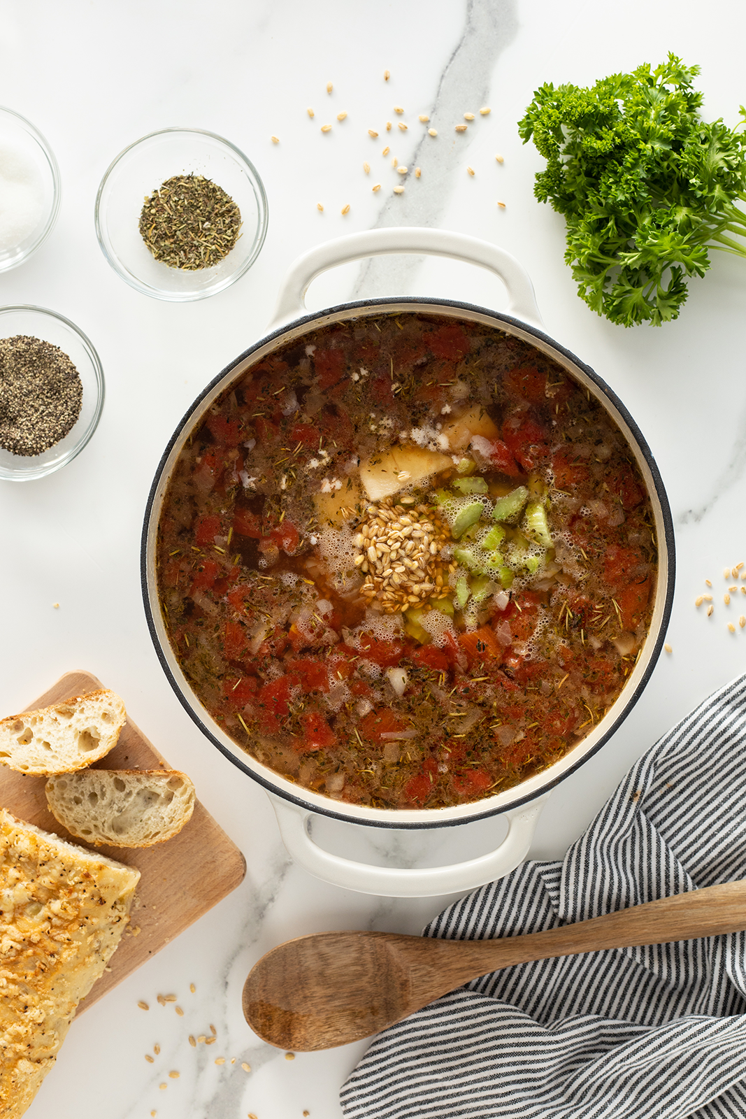 A pot of vegetable and grain soup with chopped tomatoes, celery, and herbs sits on a white counter. Nearby are a wooden spoon, striped towel, sliced bread, bowls of spices, and fresh parsley.