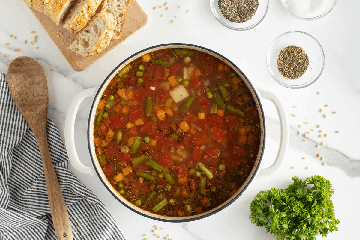 A pot of vegetable soup with diced tomatoes, potatoes, carrots, and green beans sits on a white countertop. Beside it are a wooden spoon, sliced bread, herbs, spices in bowls, and a bunch of parsley.