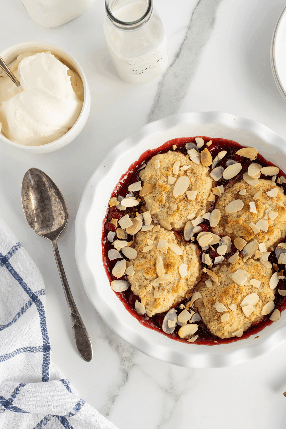 A white pie dish filled with berry cobbler topped with four biscuit rounds and sliced almonds. Nearby are a spoon, a bowl of whipped cream, glass milk bottles, and a blue-striped towel on a marble surface.