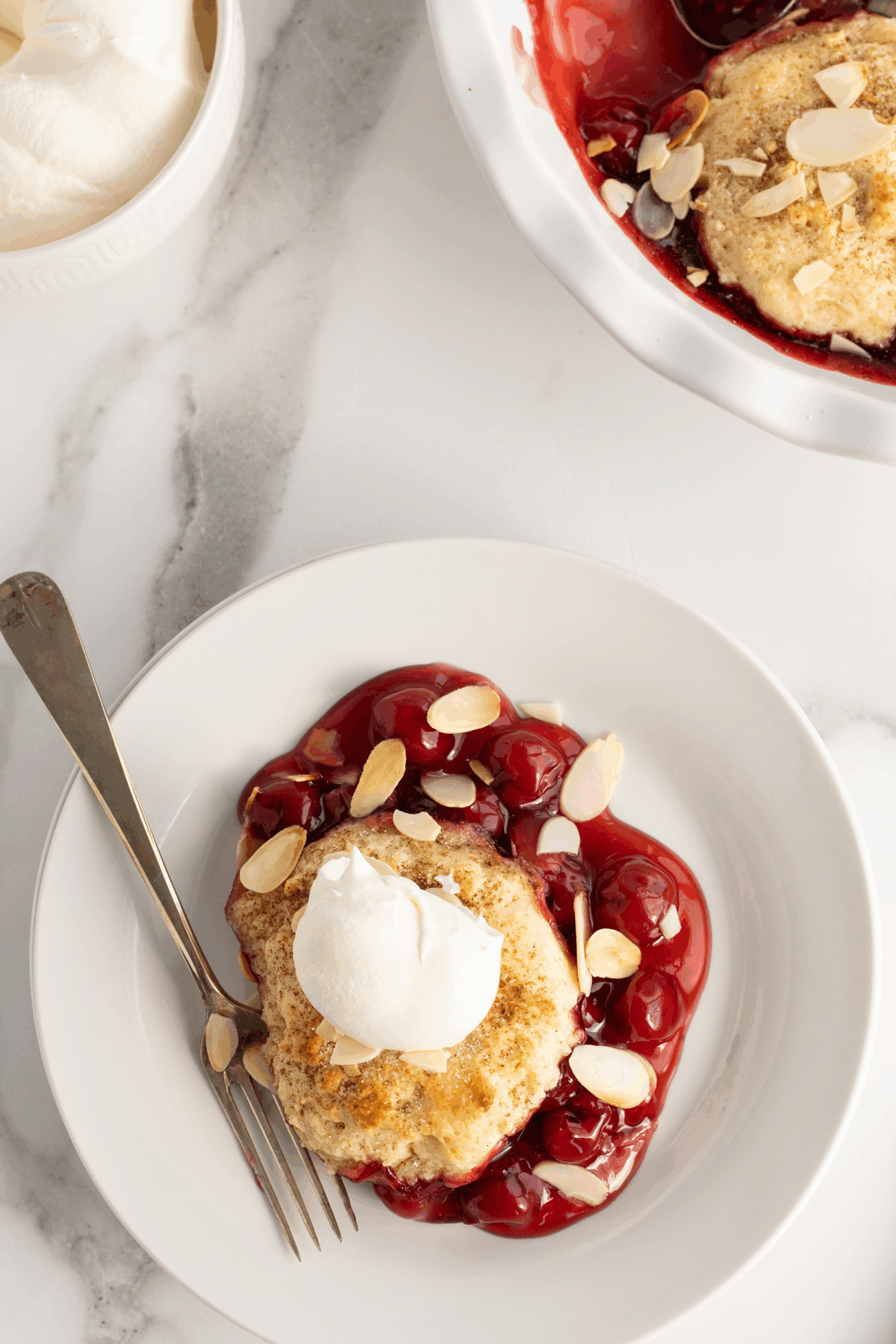 A white plate with cherry cobbler topped with a dollop of whipped cream and sliced almonds, next to a fork. A baking dish with more cobbler and a bowl of whipped cream are nearby on a marble surface.
