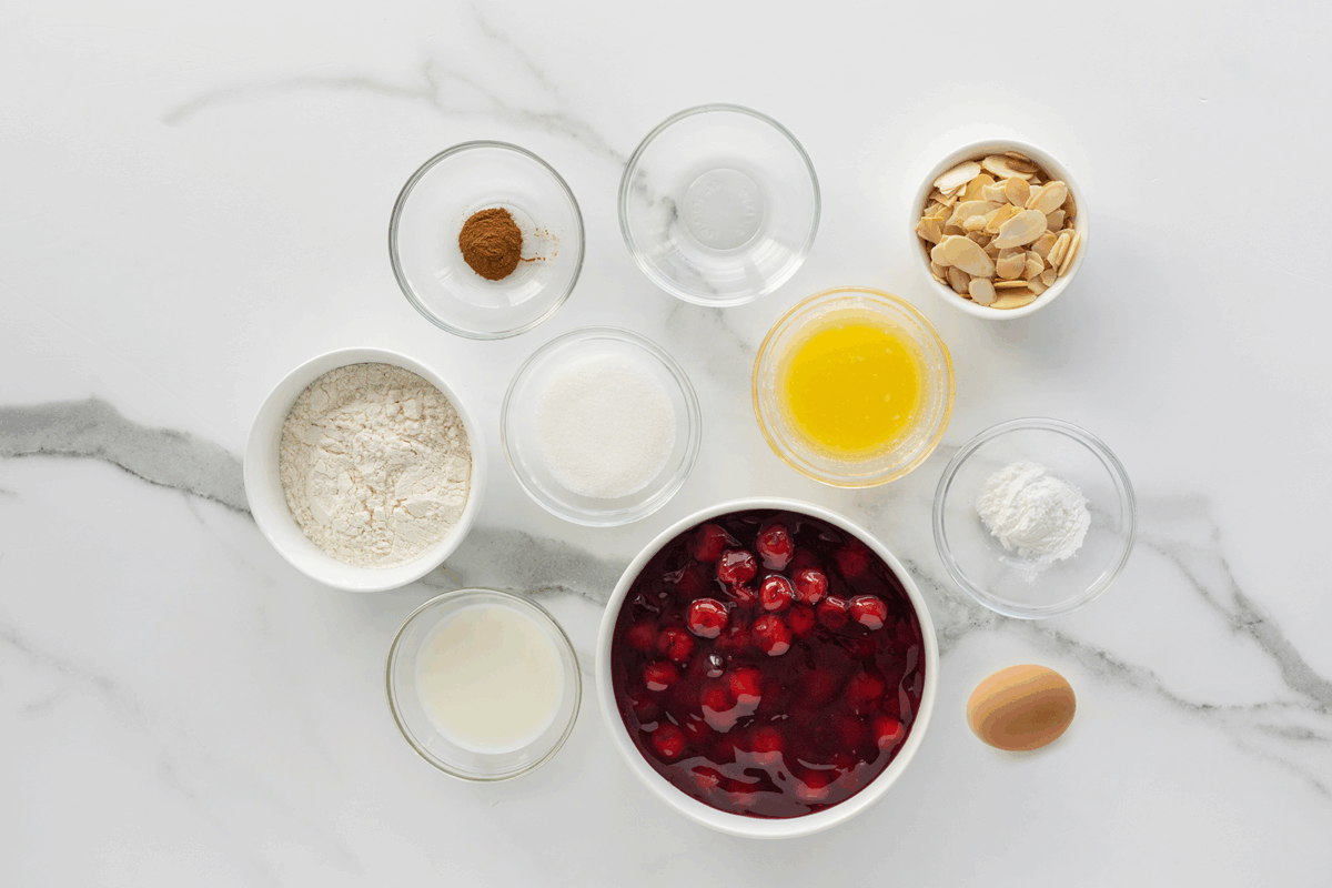A top view of baking ingredients in bowls on a marble surface, including flour, sugar, cinnamon, sliced almonds, melted butter, baking powder, milk, cherry filling, and an egg.