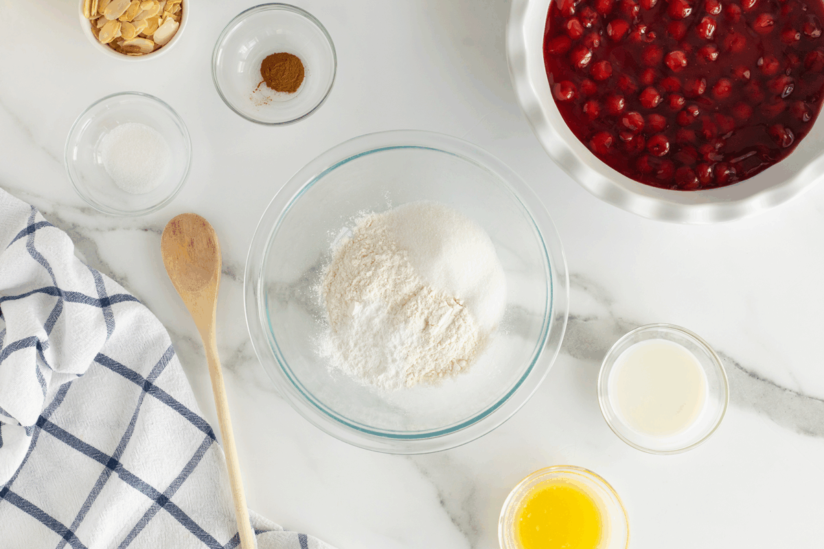 Ingredients for baking are arranged on a marble surface, including flour in a glass bowl, melted butter, milk, a bowl of cherry filling, sliced almonds, sugar, cinnamon, a wooden spoon, and a checkered kitchen towel.