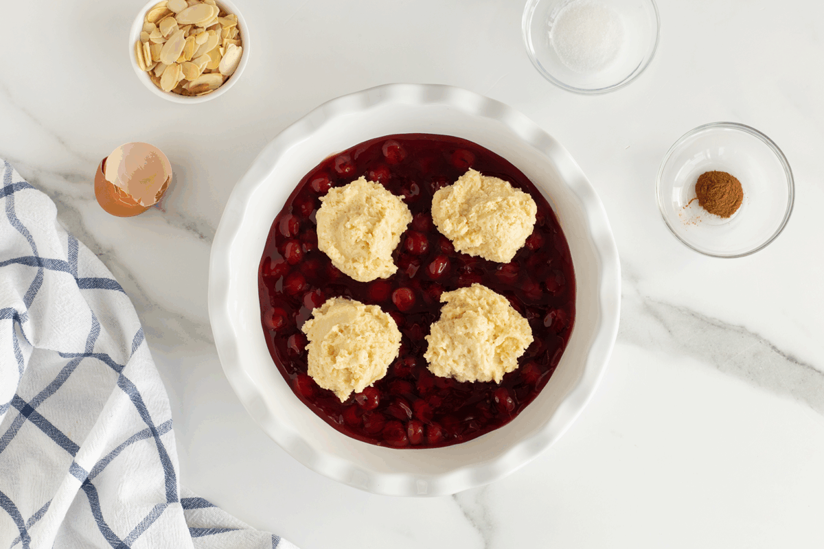 A round white baking dish filled with cherry pie filling and four dollops of biscuit dough, surrounded by small bowls of sliced almonds, sugar, cinnamon, an empty eggshell, and a blue-striped towel on a white surface.