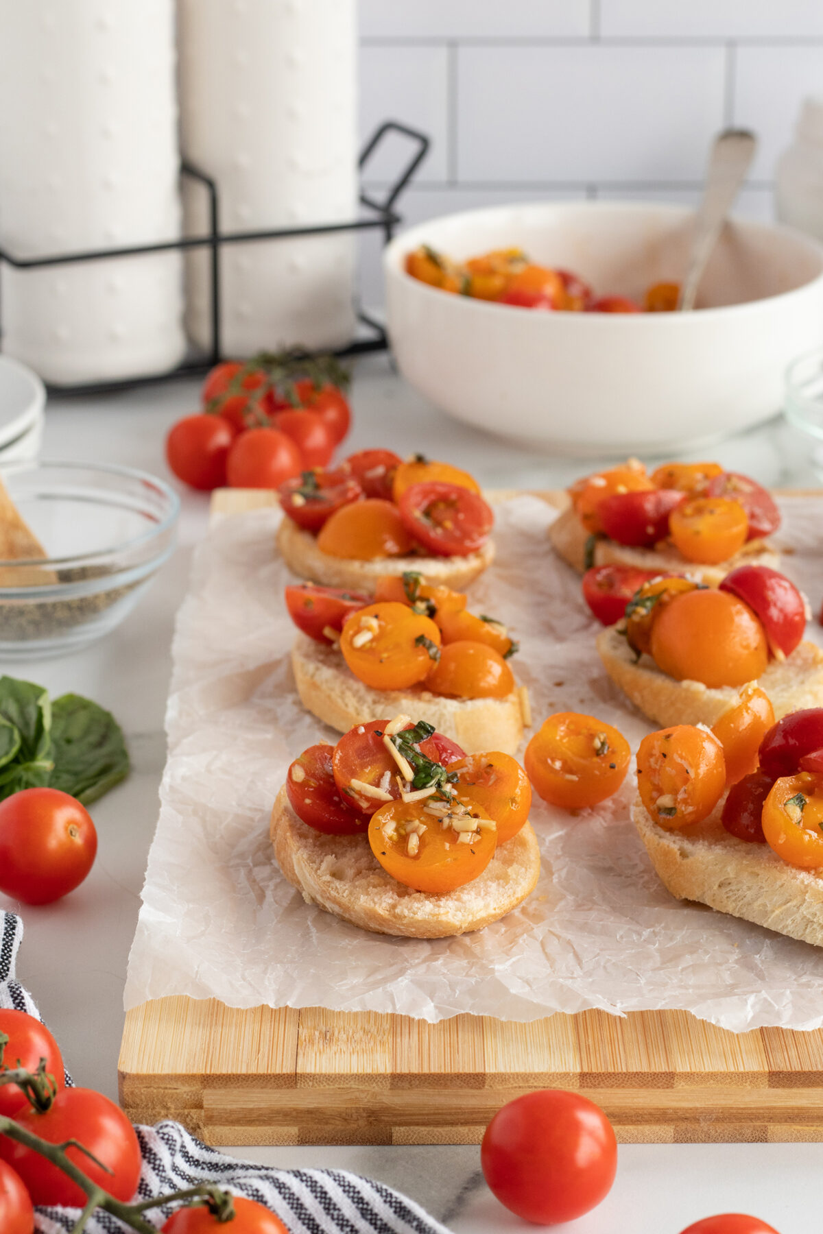 Slices of bread topped with a tomato salad made of red and yellow cherry tomatoes, herbs, and garlic, arranged on parchment paper on a wooden board. Fresh tomatoes and kitchen items are in the background.