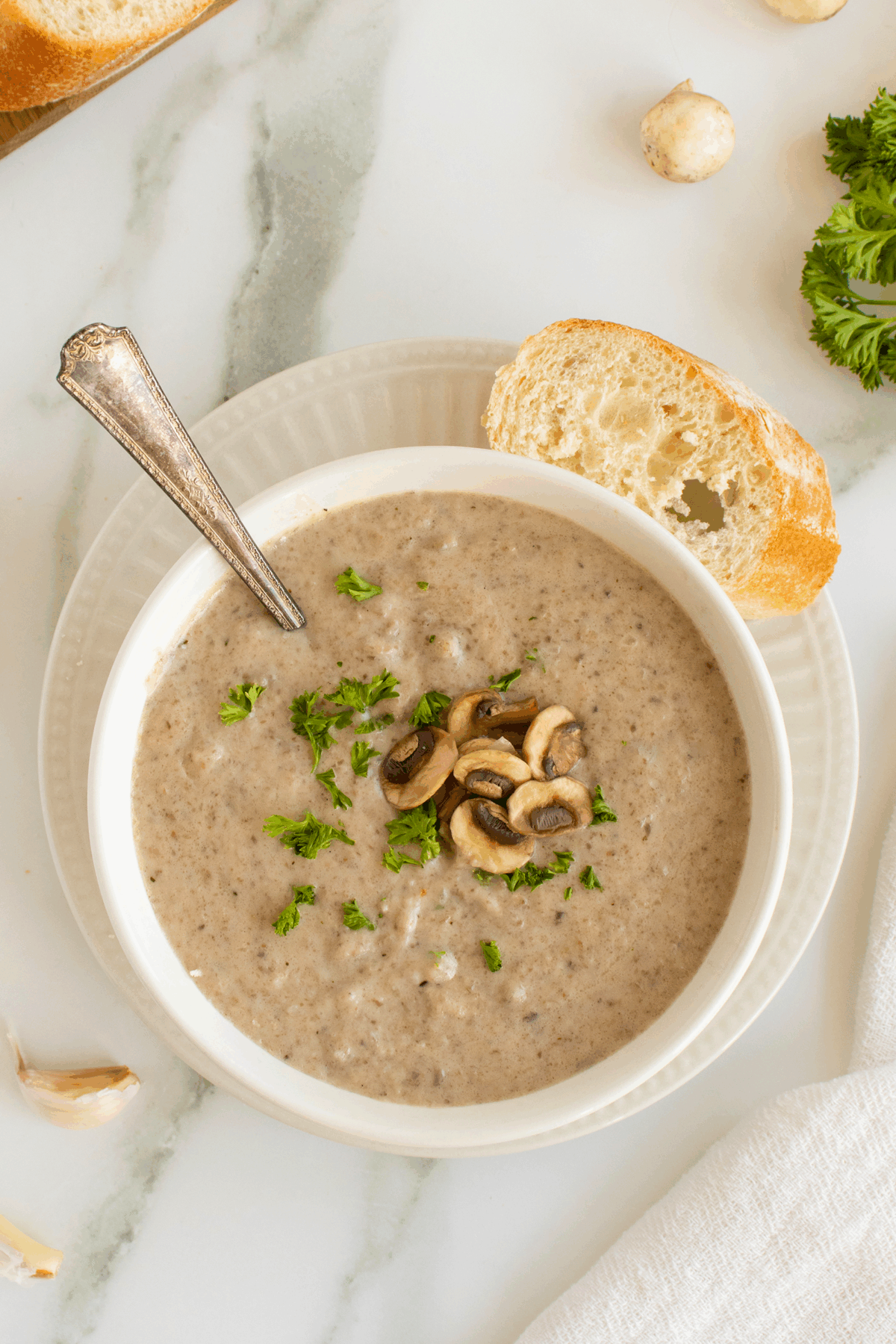 A bowl of creamy mushroom soup garnished with sliced mushrooms and chopped parsley, served with a slice of crusty bread on the side, and a silver spoon in the bowl.