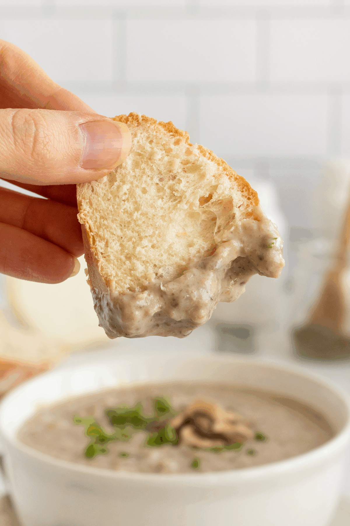 A hand holds a slice of bread dipped in creamy mushroom soup, with the bowl of soup and garnished mushrooms and herbs visible in the background.