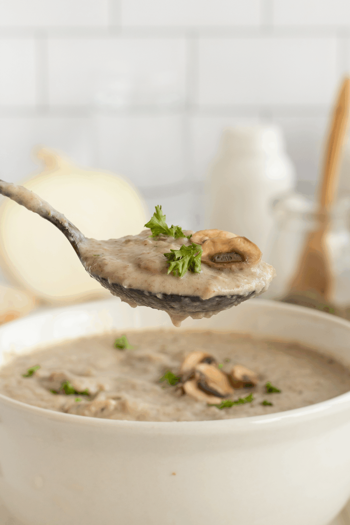 A spoonful of creamy mushroom soup garnished with chopped parsley is held above a bowl of the same soup, with mushroom slices and herbs visible. The background is softly blurred with kitchen items.