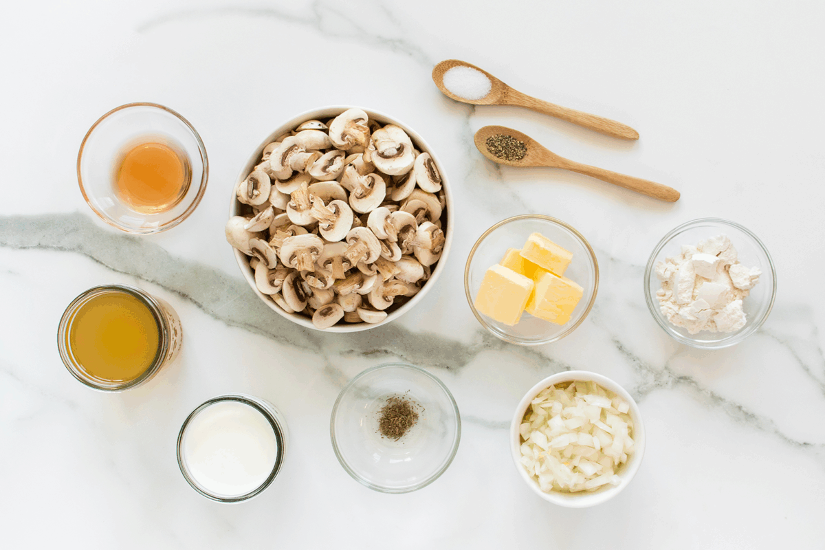 Bowls of sliced mushrooms, diced onions, butter, flour, milk, broth, vinegar, plus small bowls and spoons with salt, pepper, and dried herbs are arranged neatly on a white marble surface.